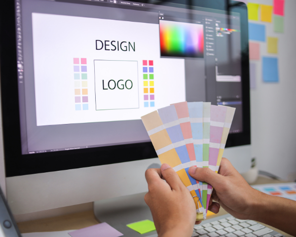 Profissional segurando amostras de cores em frente a um computador com um programa de design aberto, exibindo a criação de um logotipo e diferentes paletas de cores.